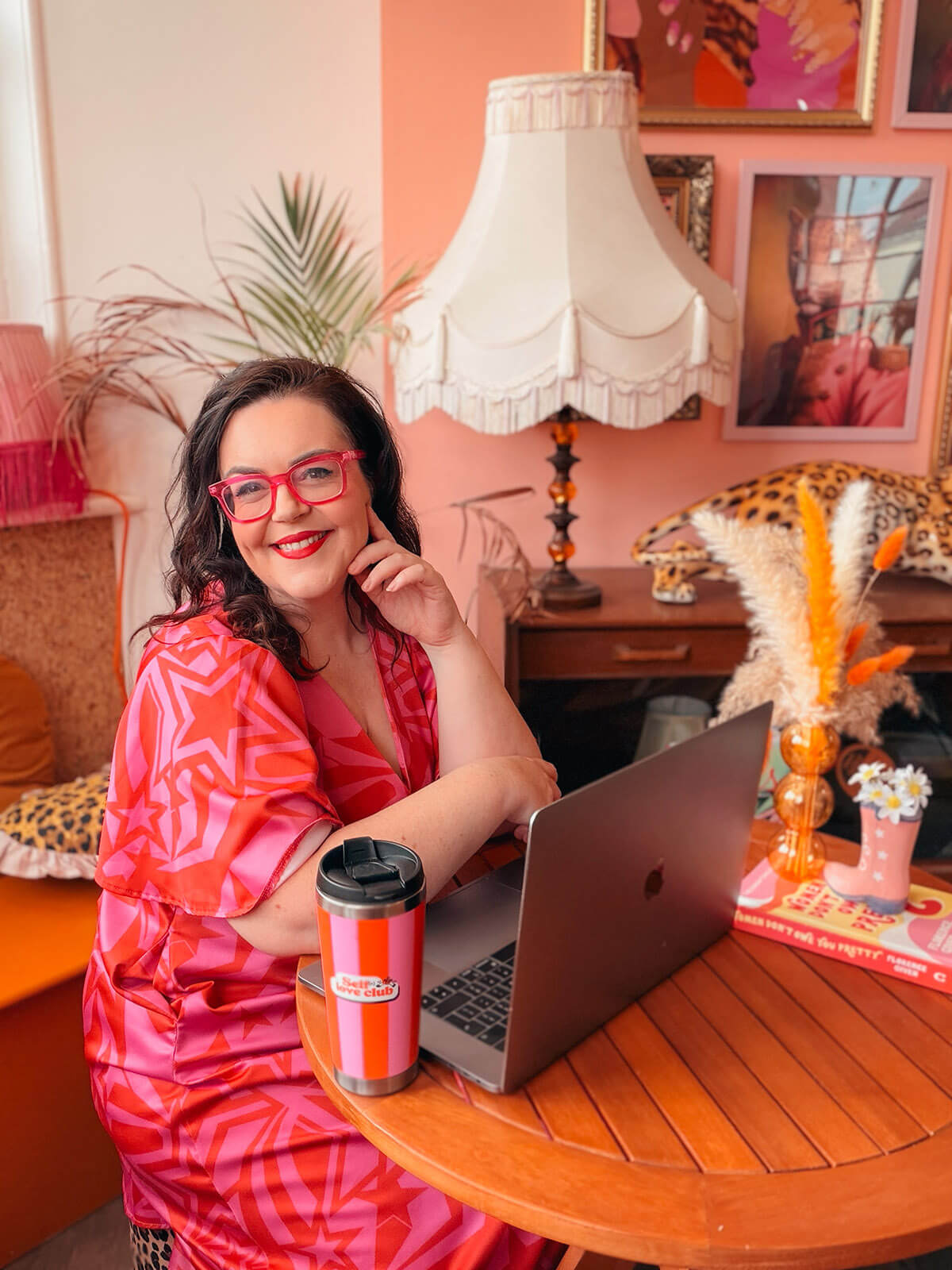Rosie Parsons smiling while working on her blog at a MacBook with a pink and orange travel mug, wearing a star-print dress and pink glasses in a vibrant pink and orange styled room