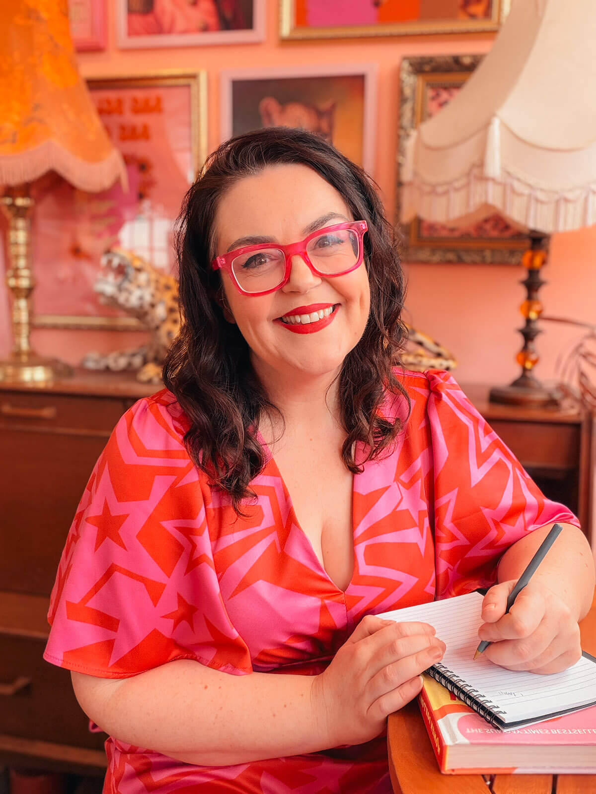 Brand photography blogger Rosie Parsons writing in a notebook, wearing a pink and red star-print dress and pink glasses in a vibrant pink styled room