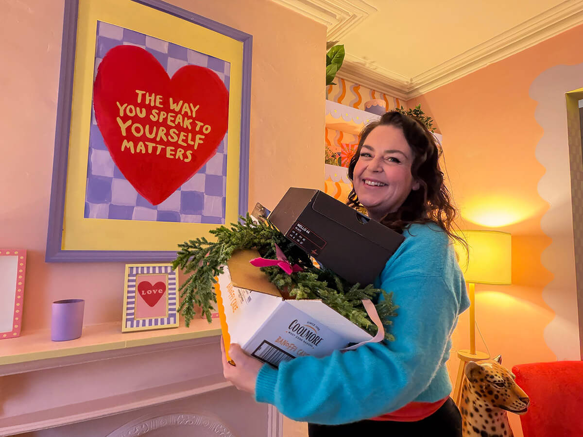 Rosie Parsons, brand photographer and business blogger, smiling while holding boxes and greenery as she declutters her home. She wears a turquoise jumper over a red top in a warm, colourful room. Behind her is a framed print of a red heart with the words "The Way You Speak to Yourself Matters." An image representing self care at home, self improvement tips and practicing self love.