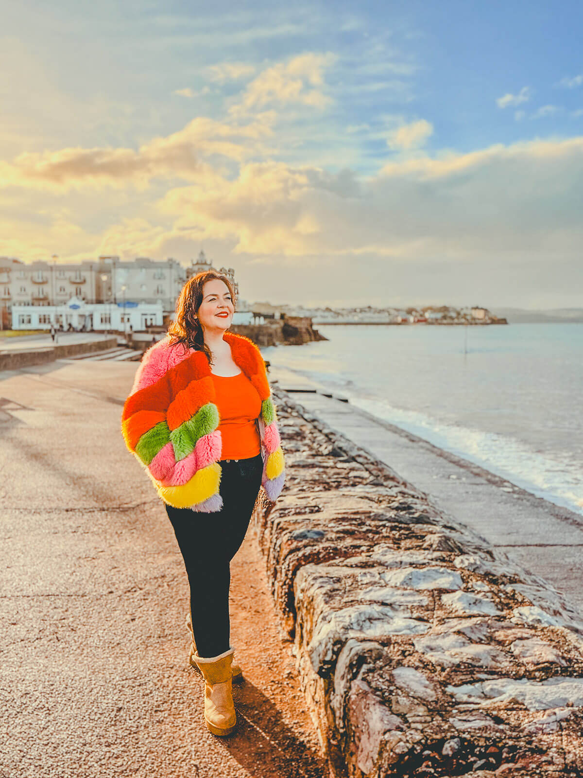 Rosie Parsons, brand photographer and business blogger, smiling while walking along a seaside promenade at golden hour. She wears a colourful faux fur jacket in pink, orange, green and yellow with an orange top, black trousers and tan boots. White coastal buildings and calm sea in the background. An uplifting image representing self care day ideas, body positivity and taking time for a healthy lifestyle.