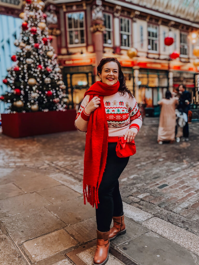Rosie Parsons, professional photographer and business blogger, smiling while holding her red scarf at Leadenhall Market in London. She wears a festive fair isle Christmas jumper with trees and gingerbread print and tan boots. A decorated Christmas tree with red and gold baubles glows behind her, showing christmas portraits at a vintage location.