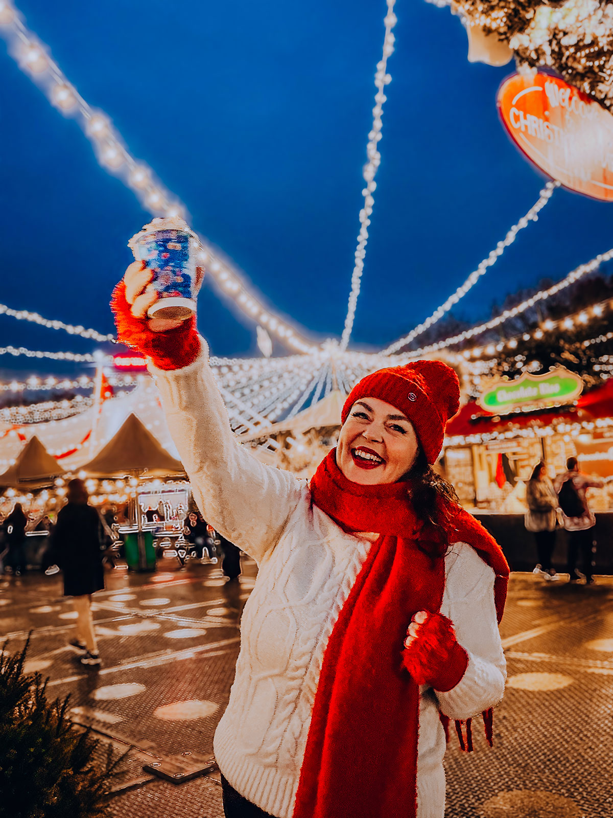 Rosie Parsons, professional photographer and business blogger, laughing while holding up a hot chocolate at a Christmas market during blue hour. She wears a cream cable-knit sweater, red bobble hat, red scarf and red gloves. String lights and market stalls glow against the evening sky, demonstrating christmas fashion photography styling.