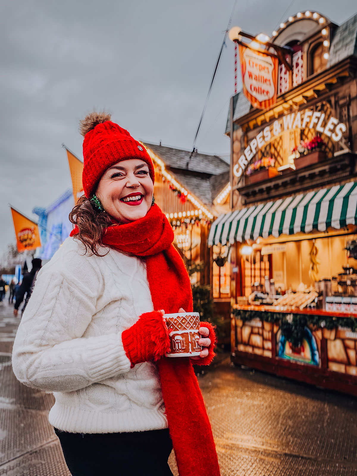 Rosie Parsons, brand photographer and business blogger, laughing while holding a gingerbread-patterned mug at a Christmas market. She wears a cream cable-knit sweater, red bobble hat, red scarf and red gloves. A wooden crepes and waffles stall glows warmly behind her, showing a cozy christmas photoshoot at a market.