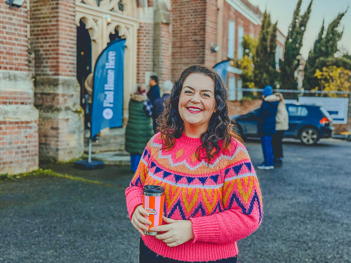 Rosie Parsons, brand photographer and business blogger, smiling outside a church holding an orange travel mug. She wears a bright pink patterned jumper with orange, yellow and navy geometric design. A historic brick building with arched doorway is in the background with people gathered nearby. An image representing selfcare tips, self improvement tips and community connection for mental and emotional health.