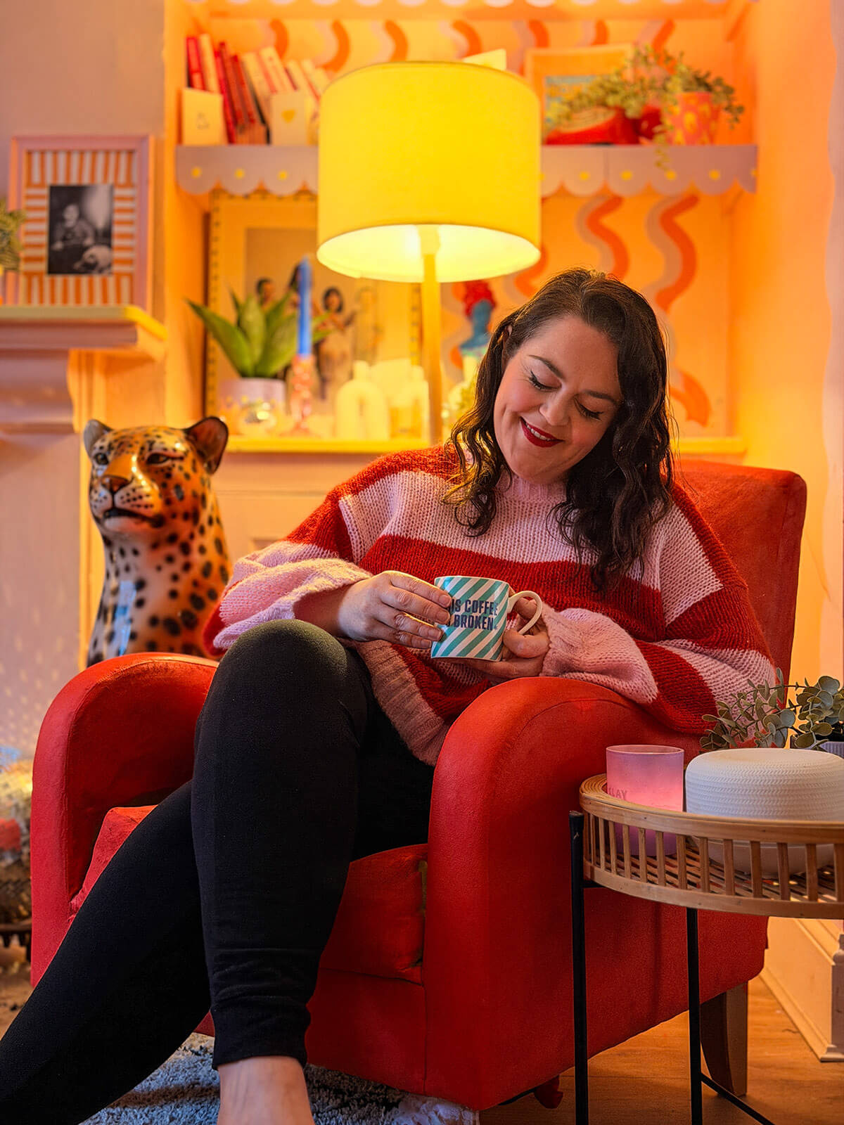 Rosie Parsons, brand photographer and business blogger, relaxing in a red velvet armchair holding a striped mug. She wears a pink and red striped jumper and smiles contentedly. The cosy room features warm lighting, a yellow lamp, colourful shelves with books and plants, and a ceramic leopard ornament. An image representing self care activities, me time and practicing self love at home.