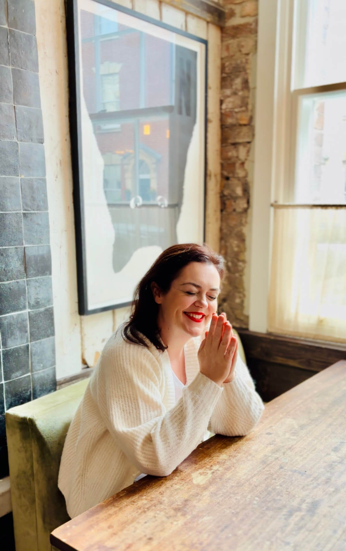 Rosie Parsons, brand photographer and business blogger, sitting peacefully at a wooden table with her hands pressed together in gratitude, eyes closed and smiling. She wears a cream knitted cardigan in a stylish cafe with exposed brick, blue tiles and natural light from a window. An image representing me time, practicing self love and selfcare ideas for mental and emotional health.