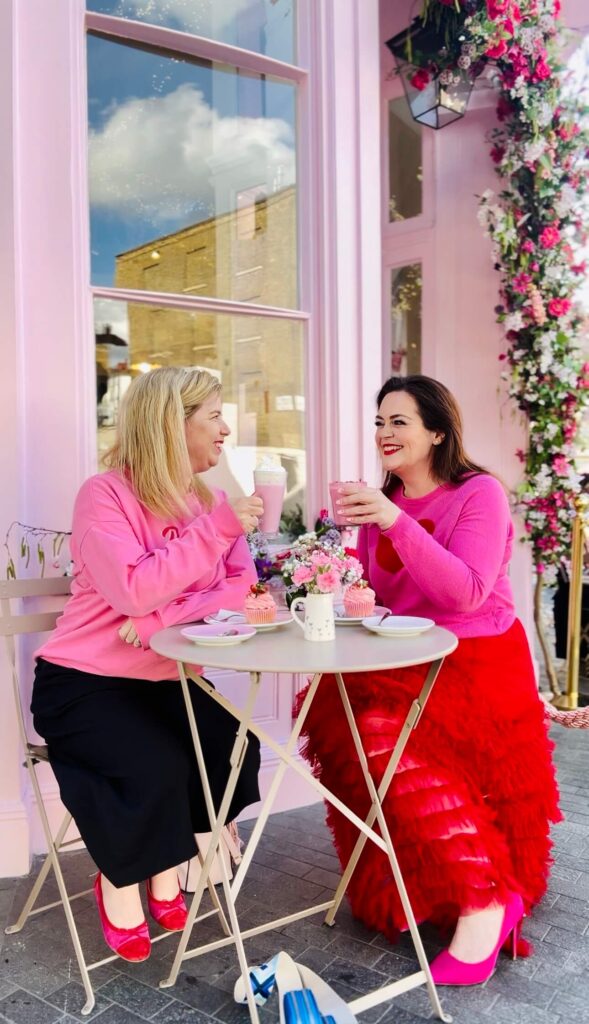 Rosie Parsons, brand photographer and business blogger, enjoying coffee and cupcakes with a friend at a pretty pink cafe with floral decorations. Rosie wears a pink top and red ruffled skirt while her blonde friend wears a pink jumper. They laugh together at a small outdoor table with pink drinks and flowers. An image representing selfcare ideas, self care day and the importance of connection for mental and emotional health.