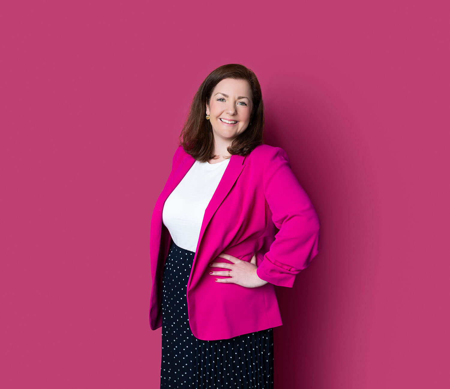 Smiling woman in a bold fuchsia blazer and white top stands confidently against a matching pink background, with a black and white polka dot skirt. Her solid-colour outfit keeps visual focus on her face, illustrating why clean, bold tones work better than busy patterns in headshots.