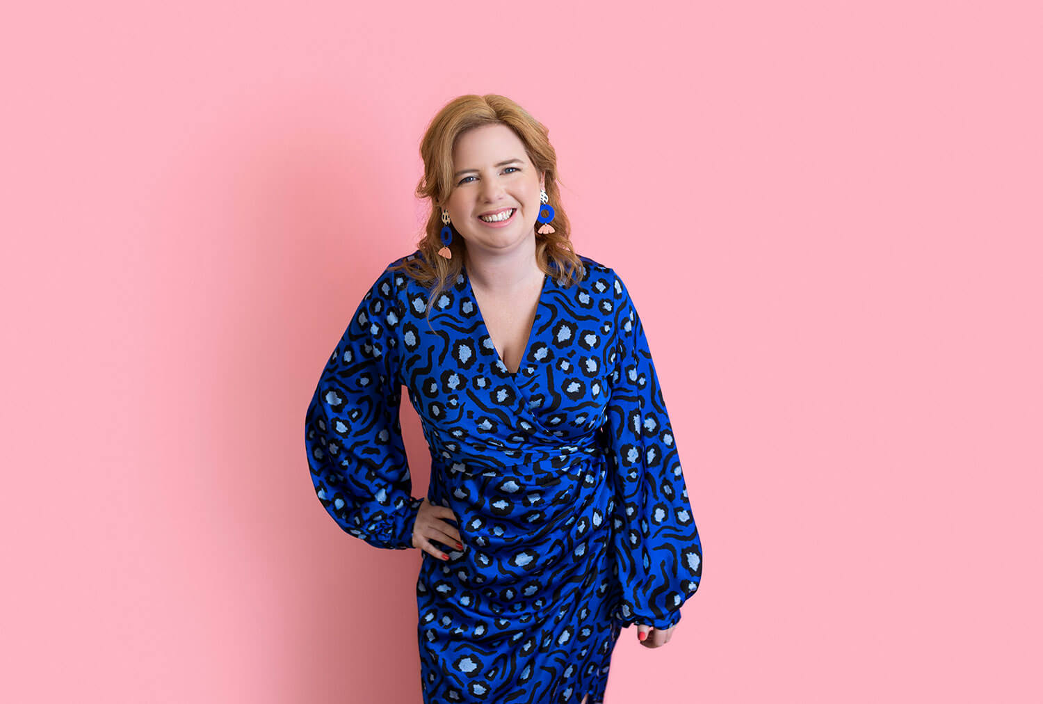 Smiling woman in a blue patterned dress and statement earrings poses confidently against a soft pink background. Her bold accessories and vibrant outfit reflect what to wear for headshots with a creative personal brand, showing how colour and shape photograph well on camera.