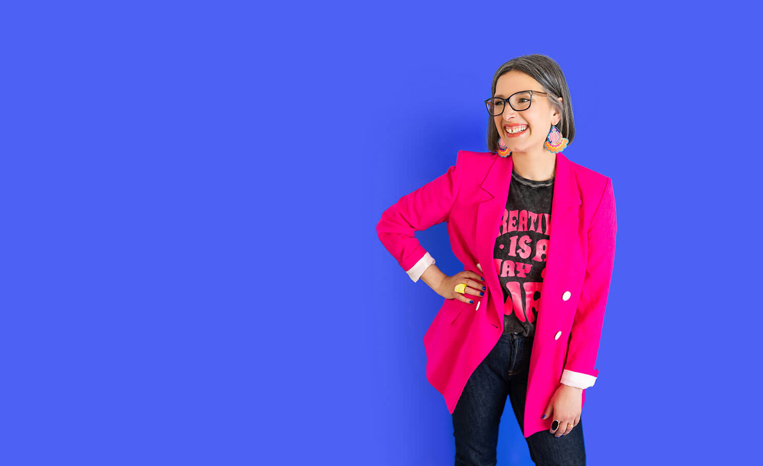 Woman smiling confidently in a bright pink blazer, graphic tee that reads “Creativity is a way of life,” and dark jeans, standing against a solid blue background. Her bold outfit reflects a creative, personal brand style, illustrating what to wear for headshots in a non-corporate industry.
