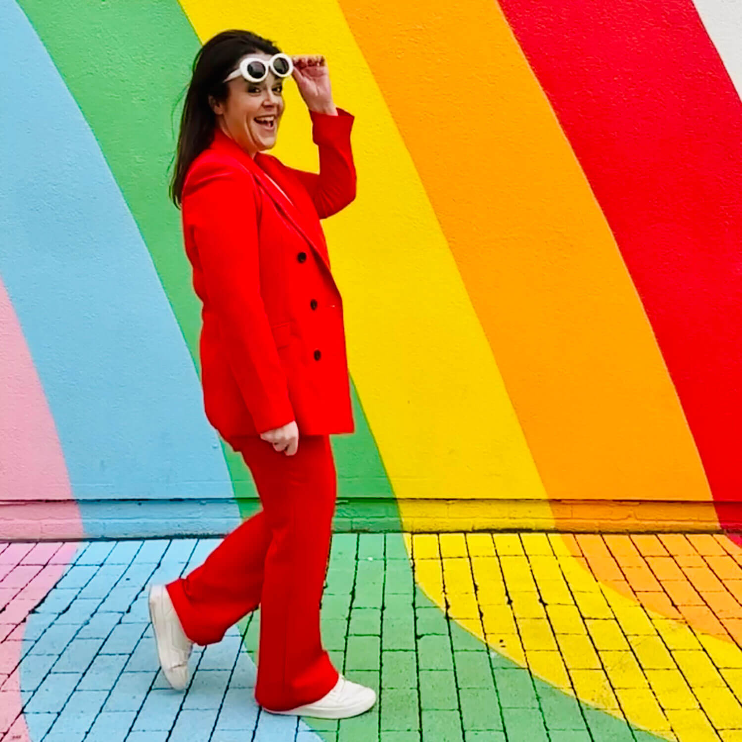 Brand photographer Rosie Parsons walks confidently past a rainbow mural, smiling playfully in a bright red suit and white sunglasses. This self-portrait captures natural movement, showing how to take good pictures of yourself by replacing stiff poses with simple actions.