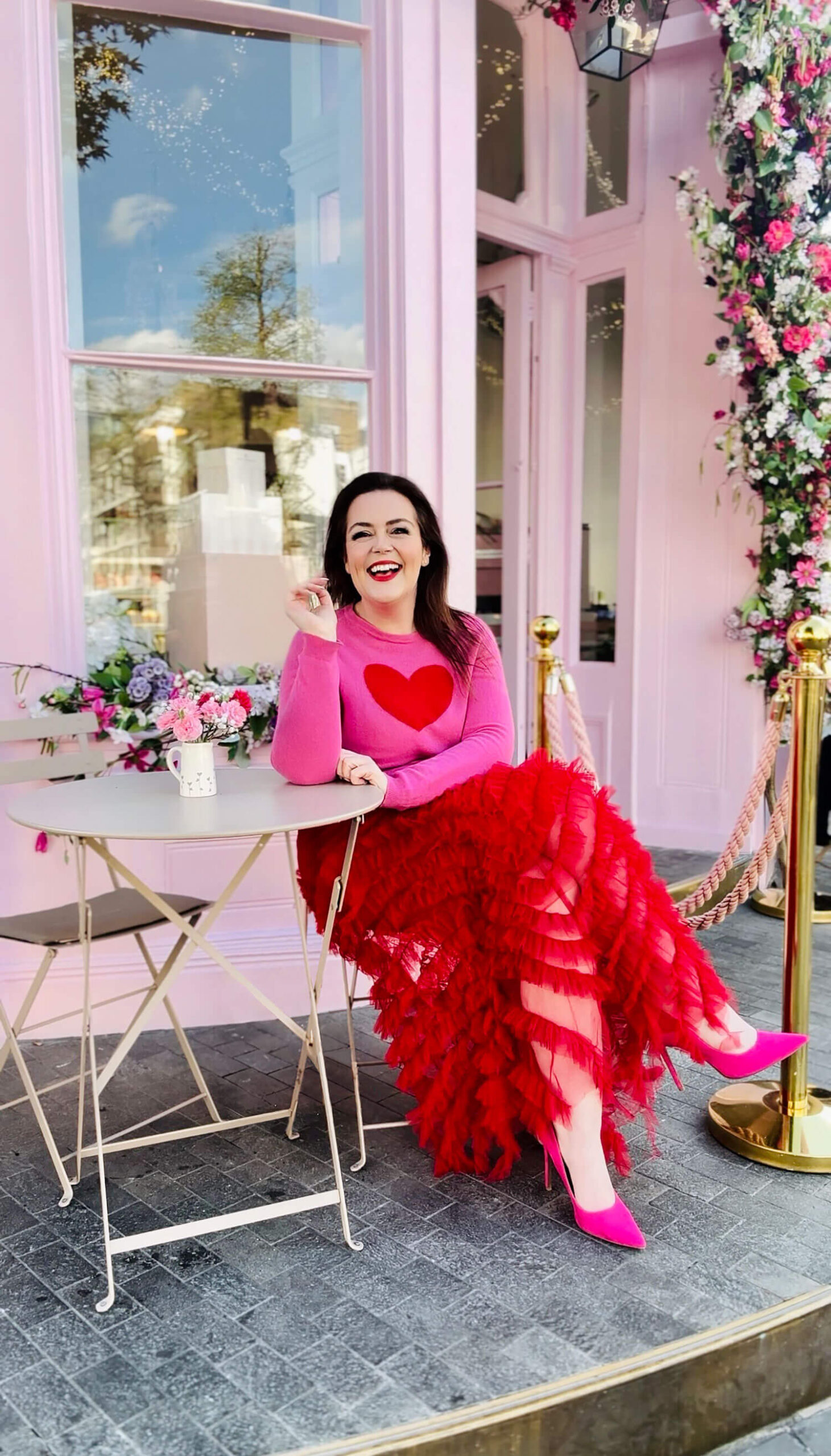 Brand photographer Rosie Parsons sits at an outdoor café table in a bright pink and red outfit, smiling confidently in front of a pastel pink storefront decorated with flowers. This vibrant self-portrait shows how to take good pictures of yourself for personal branding and profile photos by using color, location, and authentic expression.