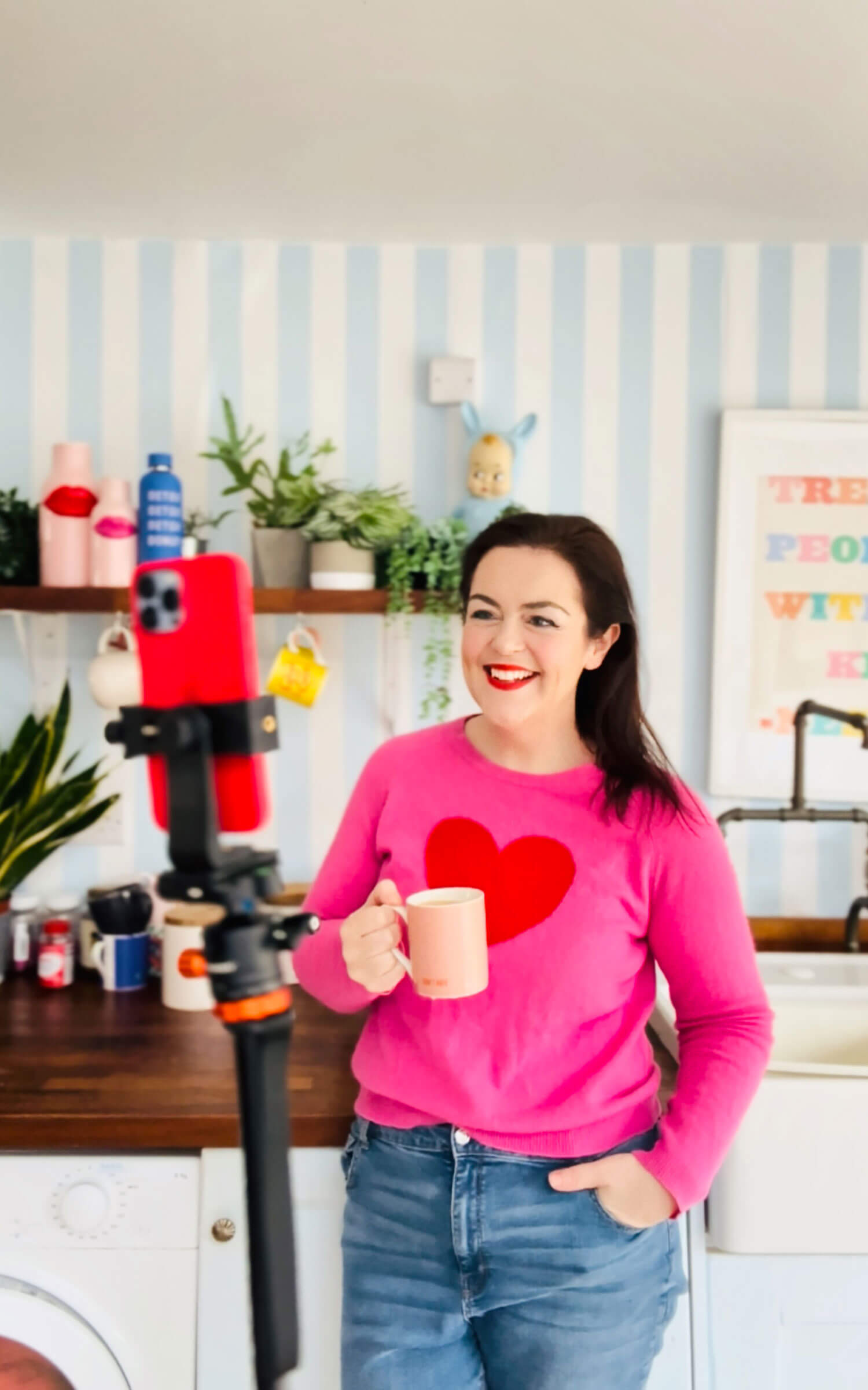 Brand photographer Rosie Parsons smiles at a phone mounted on a tripod while holding a mug in her kitchen, wearing a pink sweater with a red heart. This home setup shows how to take good pictures of yourself using intentional distance, natural lighting, and a relaxed posture.